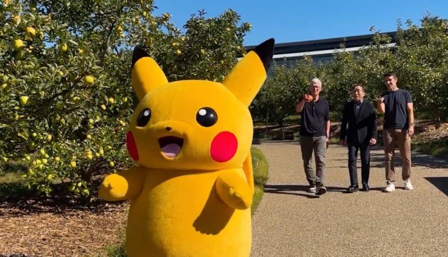 Eine Person im Pikachu-Kostüm steht im Vordergrund eines Apfelgartens, während drei Männer im Hintergrund auf einem Weg spazieren.